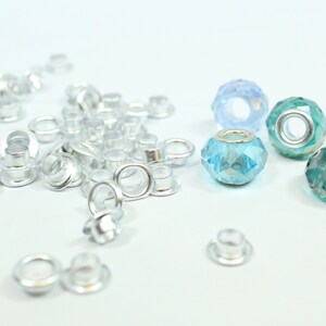 May include: A close-up of silver metal eyelets and blue and green glass beads. The eyelets are scattered on a white surface. The beads are arranged in a row.