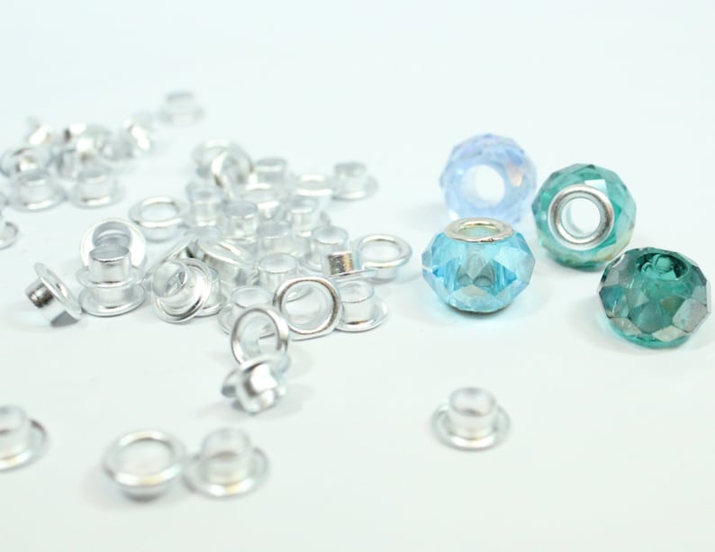 May include: A close-up of silver metal eyelets and blue and green glass beads. The eyelets are scattered on a white surface. The beads are arranged in a row.