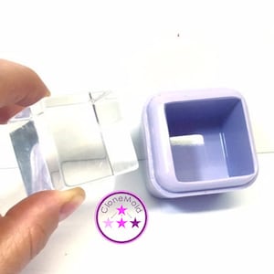 Può includere: Uno stampo quadrato trasparente tenuto in mano, accanto a uno stampo quadrato color lavanda. Lo stampo trasparente è trasparente, mentre l'altro ha un'apertura quadrata. È visibile un logo circolare con stelle.