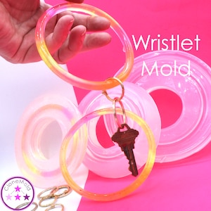 May include: A clear, round wristlet mold with a key attached. The wristlet is a gradient of yellow and orange. The text "Wristlet Mold" is in white. The background is pink.