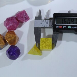 May include: A set of six colorful dice with glitter, measured with a digital caliper showing 16.09 mm. The dice are in various shapes, including a pyramid, a cube, and a diamond.