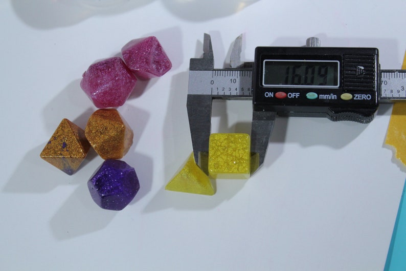 May include: A set of six colorful dice with glitter, measured with a digital caliper showing 16.09 mm. The dice are in various shapes, including a pyramid, a cube, and a diamond.