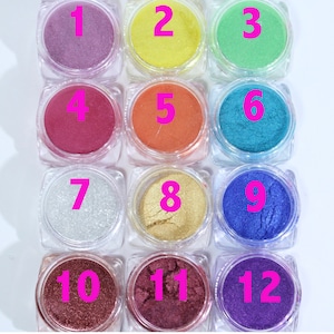 Puede incluir: Una colecci&oacute;n de doce peque&ntilde;os frascos transparentes llenos de polvos cosm&eacute;ticos coloridos. Los polvos var&iacute;an en color, desde lavanda hasta dorado y morado oscuro, con cada frasco numerado en rosa.