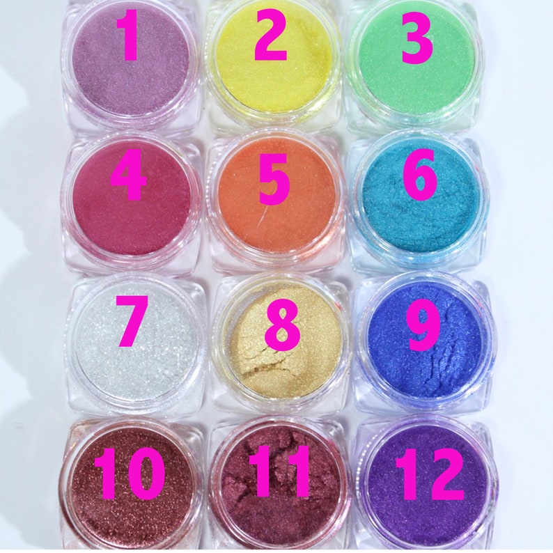 Puede incluir: Una colecci&oacute;n de doce peque&ntilde;os frascos transparentes llenos de polvos cosm&eacute;ticos coloridos. Los polvos var&iacute;an en color, desde lavanda hasta dorado y morado oscuro, con cada frasco numerado en rosa.