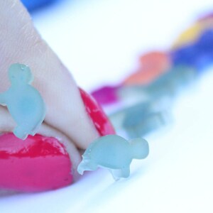 May include: Two small, translucent blue turtle-shaped charms are held by a person with red nail polish. A row of colorful charms in various colors, including blue, orange, and green, are blurred in the background. The charms appear to be made of a soft material.