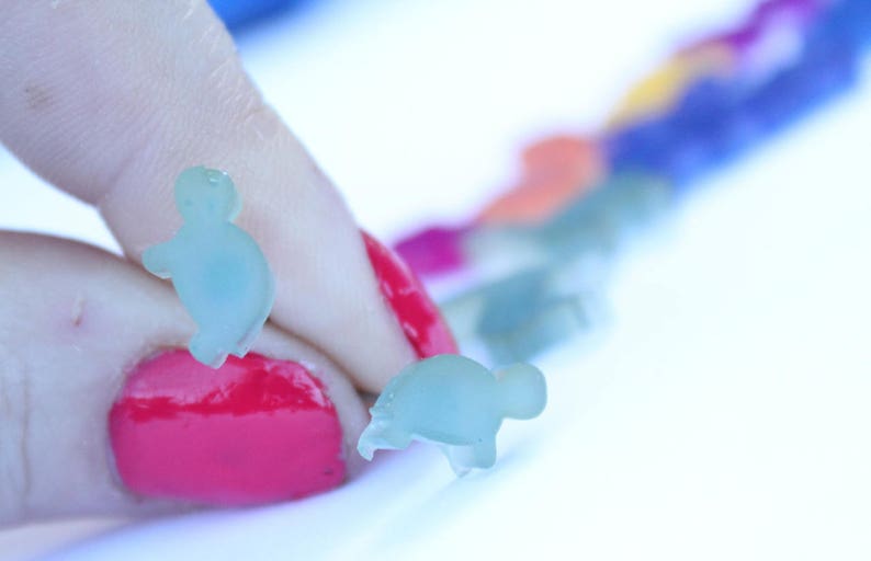 May include: Two small, translucent blue turtle-shaped charms are held by a person with red nail polish. A row of colorful charms in various colors, including blue, orange, and green, are blurred in the background. The charms appear to be made of a soft material.
