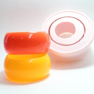 May include: Two stacked plastic bangles, one orange and one yellow. The bangles are smooth and glossy. A pink silicone mould for making bangles is in the background.