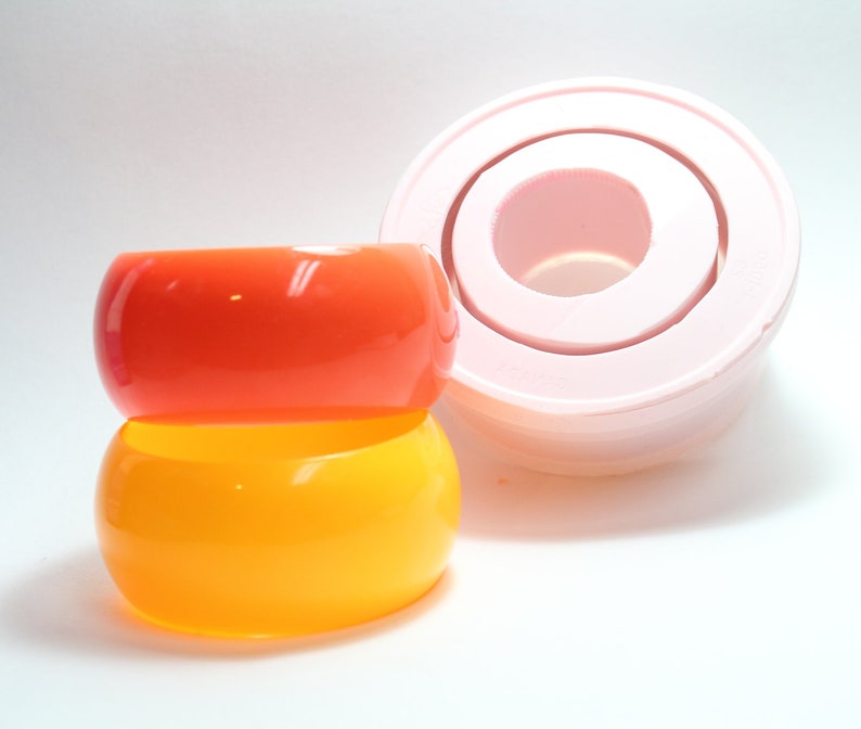 May include: Two stacked plastic bangles, one orange and one yellow. The bangles are smooth and glossy. A pink silicone mould for making bangles is in the background.