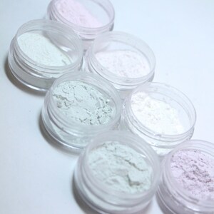 May include: A collection of small, clear plastic jars filled with various pastel-colored powders. The powders include shades of pink, white, and green. The jars are arranged in a diagonal pattern, showcasing the different colors and textures.