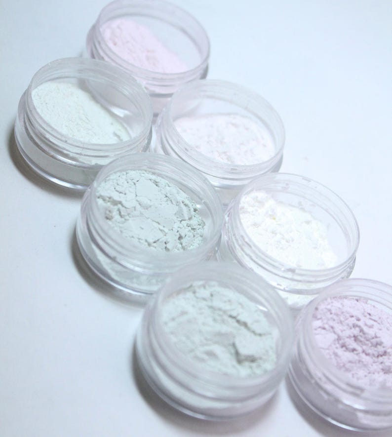 May include: A collection of small, clear plastic jars filled with various pastel-colored powders. The powders include shades of pink, white, and green. The jars are arranged in a diagonal pattern, showcasing the different colors and textures.