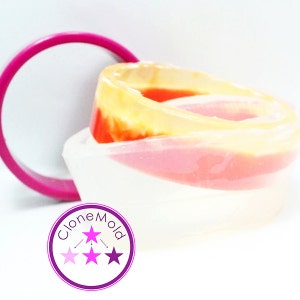 May include: A layered resin mold with a pink, orange, and yellow design. A bright pink circular object is next to the mold. The mold is in a white container. The text "CloneMold" is in a purple circle with stars.