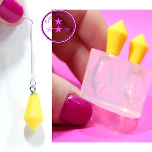 May include: A single earring with a silver chain and a bright yellow, geometric charm. The image also shows a silicone mould with two diamond-shaped cavities and yellow inserts, used for crafting jewellery. The background is a vibrant pink.