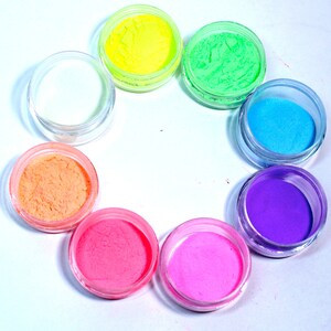 May include: Eight small jars of colorful glitter powder arranged in a circle on a white surface. The colors are yellow, green, blue, purple, pink, orange, and white.