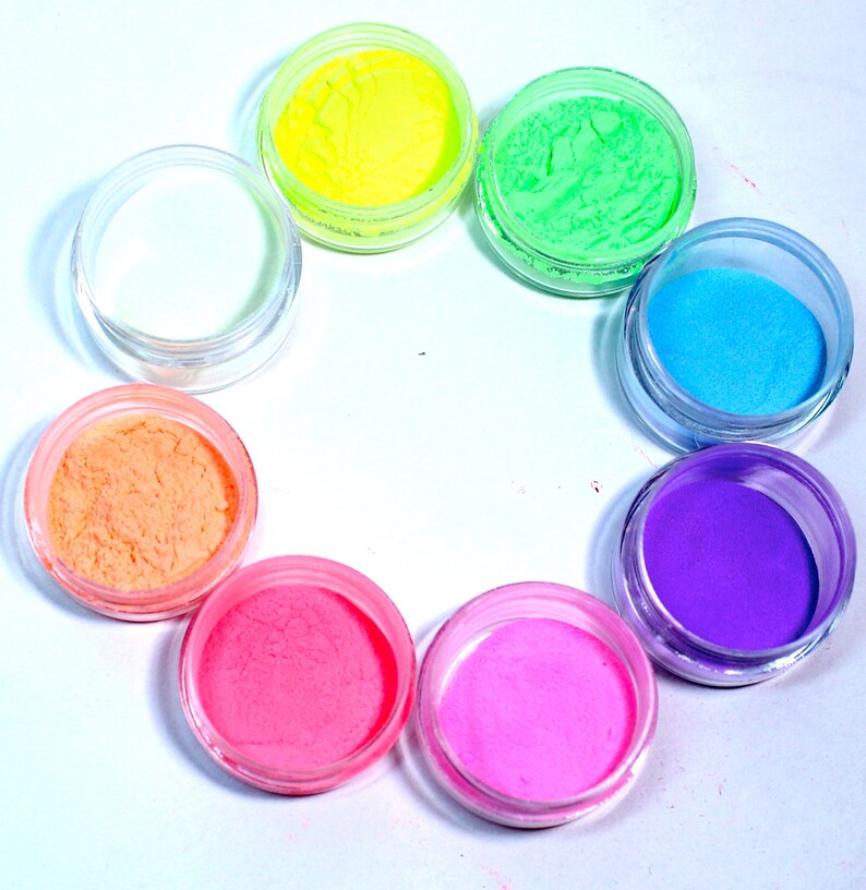 May include: Eight small jars of colorful glitter powder arranged in a circle on a white surface. The colors are yellow, green, blue, purple, pink, orange, and white.