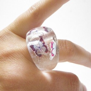 May include: A clear glass ring with a chunky design features pink and white butterflies embedded inside. The ring is worn on a finger.