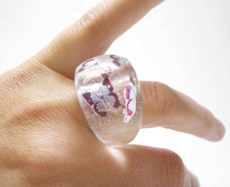 May include: A clear glass ring with a chunky design features pink and white butterflies embedded inside. The ring is worn on a finger.