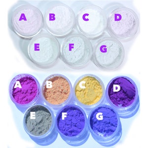May include: An overhead shot displays fourteen small, clear plastic jars filled with colorful powders. The top row features pastel shades, while the bottom row showcases vibrant hues, including pink, orange, yellow, and purple. Each jar is labeled with a letter.