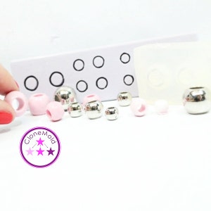 May include: An assortment of beads in various sizes and colors, including pink and silver. The beads are displayed with a white mold and a card with black circles. The image also includes the logo "CloneMold".