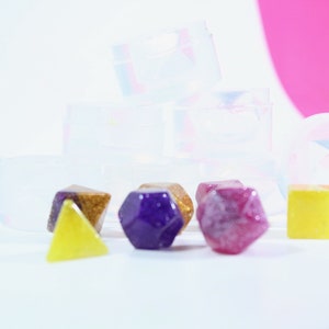 May include: A set of six colorful resin dice in various shapes, including a purple hexagonal die, a red diamond-shaped die, a yellow triangular die, and a gold and purple die. The dice are sitting on a white surface in front of several clear plastic containers.