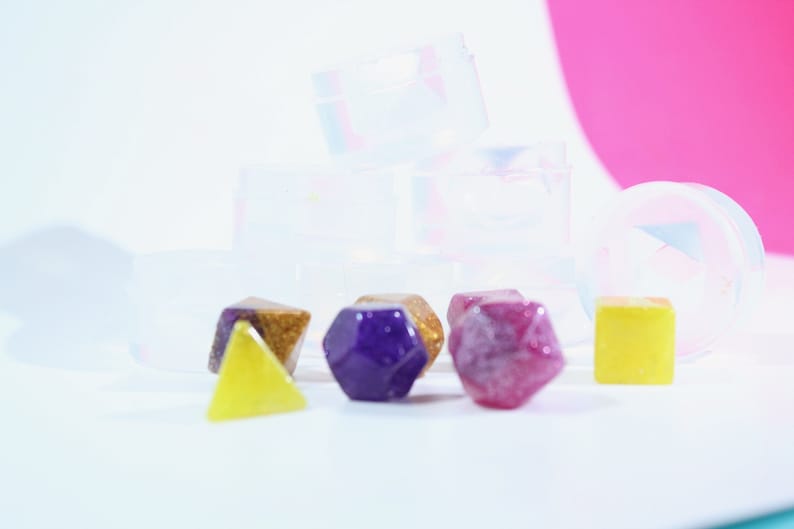 May include: A set of six colorful resin dice in various shapes, including a purple hexagonal die, a red diamond-shaped die, a yellow triangular die, and a gold and purple die. The dice are sitting on a white surface in front of several clear plastic containers.