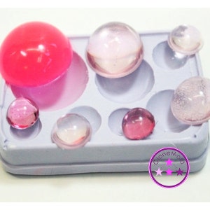 May include: A gray silicone mold filled with various sizes of clear and pink resin spheres. The spheres range in color from clear to deep pink. The mold has several circular indentations to hold the spheres. The image includes a logo in the bottom right corner.