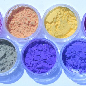 May include: Seven small, clear plastic jars filled with colorful powders. The colors include shades of pink, orange, yellow, purple, blue, and green. The powders appear finely milled and are arranged in a circular pattern.