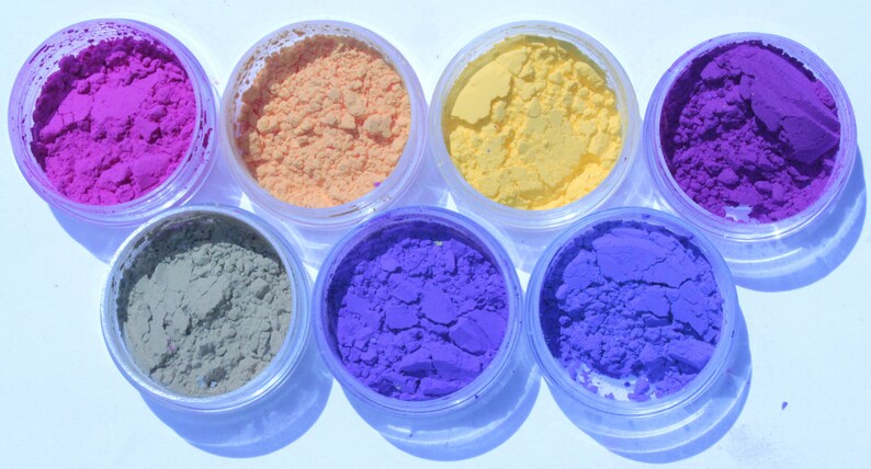 May include: Seven small, clear plastic jars filled with colorful powders. The colors include shades of pink, orange, yellow, purple, blue, and green. The powders appear finely milled and are arranged in a circular pattern.