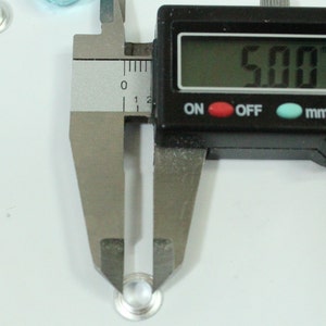 May include: A close-up of a silver metal rivet with a digital caliper measuring 5.00 mm. The caliper is black with a digital display and buttons labeled "ON", "OFF", and "mm/in".