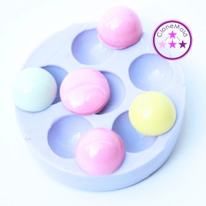 May include: A light purple silicone mold filled with six colorful, semi-spherical candies. The candies are pastel pink, yellow, and light blue. The image includes a logo in the upper right corner.