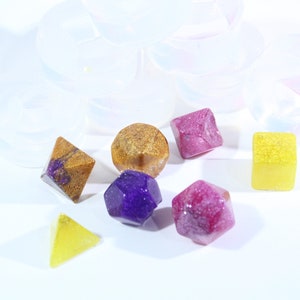 May include: A set of six colorful resin dice with glitter embedded in the resin. The dice are in various shapes, including a cube, a pyramid, a dodecahedron, and a tetrahedron. The dice are a mix of purple, pink, yellow, and gold.
