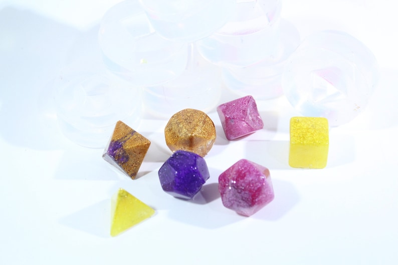 May include: A set of six colorful resin dice with glitter embedded in the resin. The dice are in various shapes, including a cube, a pyramid, a dodecahedron, and a tetrahedron. The dice are a mix of purple, pink, yellow, and gold.
