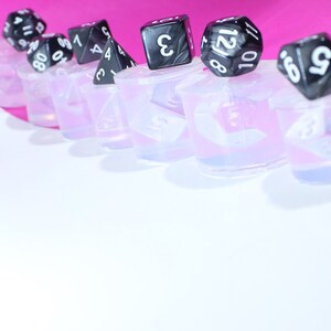 May include: A row of clear silicone molds with black dice on top. The dice have white numbers on them. The molds are on a pink surface.