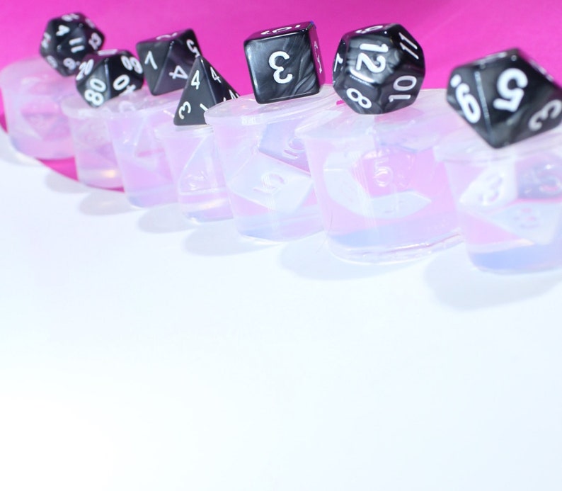 May include: A row of clear silicone molds with black dice on top. The dice have white numbers on them. The molds are on a pink surface.