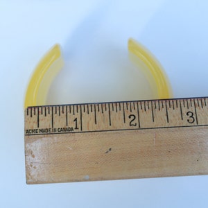 May include: A translucent yellow bangle bracelet, approximately 3 inches in diameter, is shown next to a wooden ruler. The bracelet has a curved, smooth design. The ruler is marked with black lines and numbers, and the text "ACME MADE IN CANADA" is visible.