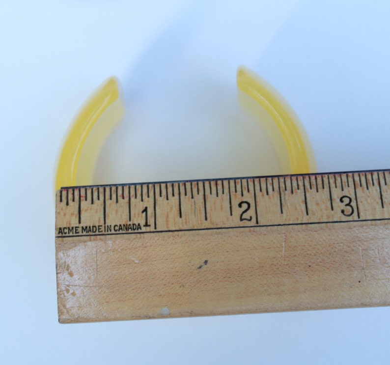 May include: A translucent yellow bangle bracelet, approximately 3 inches in diameter, is shown next to a wooden ruler. The bracelet has a curved, smooth design. The ruler is marked with black lines and numbers, and the text "ACME MADE IN CANADA" is visible.