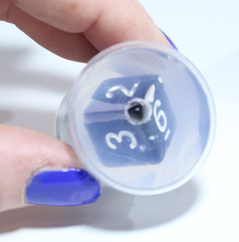 May include: A blue six-sided die with white numbers 2, 3, 5, and 6 visible. The die is inside a clear plastic container.