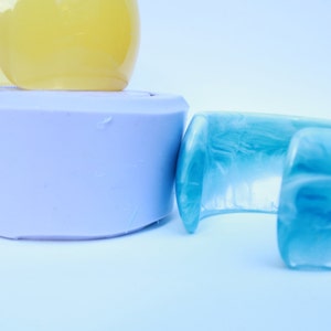 May include: A collection of resin jewelry and accessories. The image features a light blue cuff bracelet with a marbled effect, a lavender-colored cylindrical object, and two yellow, rounded objects. The items are set against a white background.
