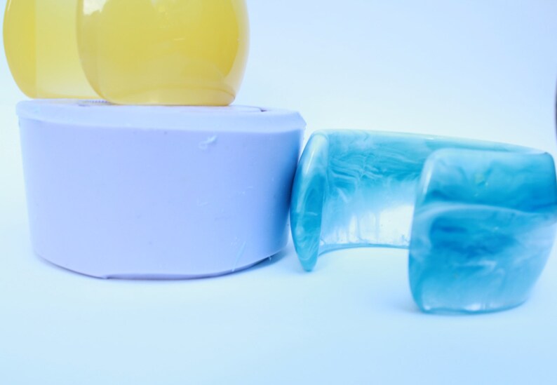 May include: A collection of resin jewelry and accessories. The image features a light blue cuff bracelet with a marbled effect, a lavender-colored cylindrical object, and two yellow, rounded objects. The items are set against a white background.