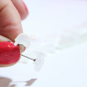 May include: Close-up of clear, plastic, animal-shaped push pins. One pin is being held by a hand with red nail polish. The pins are on a white surface. The pins are small and translucent.