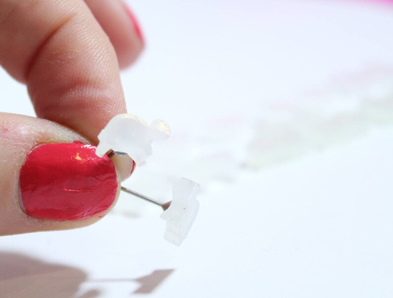 May include: Close-up of clear, plastic, animal-shaped push pins. One pin is being held by a hand with red nail polish. The pins are on a white surface. The pins are small and translucent.