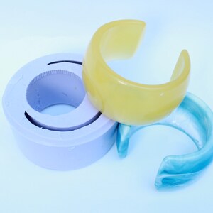 May include: A light yellow and a light blue cuff bracelet are displayed next to a purple mold. The bracelets have a curved, open design. The mold is circular with a ridged interior.