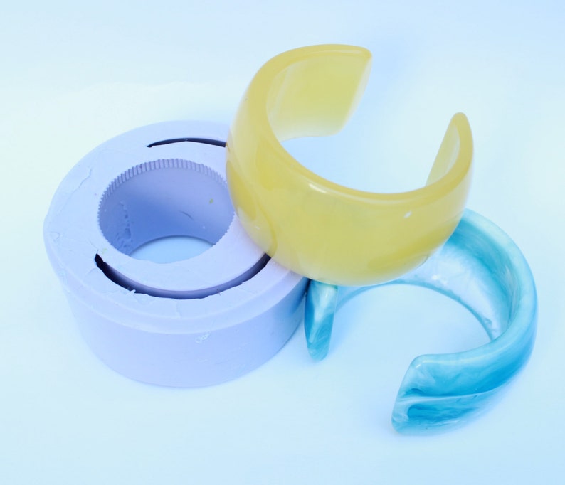May include: A light yellow and a light blue cuff bracelet are displayed next to a purple mold. The bracelets have a curved, open design. The mold is circular with a ridged interior.