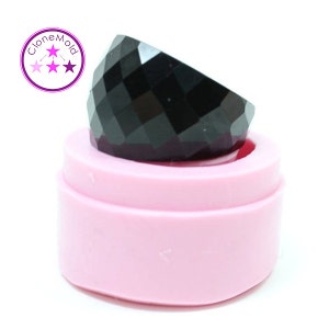 May include: A faceted black gemstone sits inside a light pink silicone mold. The gemstone has a geometric design. The mold is round with a two-tiered design. The image includes a logo with the text "CloneMold".