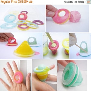 K&ouml;nnte beinhalten: Ein Set aus bunten Harzringen mit verschiedenen Designs, darunter ein Ring mit einer Kuppelform und ein Ring mit einer flachen Oberseite. Die Ringe sind aus klarem Harz gefertigt und haben verschiedene Farben, darunter Pink, Gelb, Blau und Gr&uuml;n. Die Ringe werden auf einem wei&szlig;en Hintergrund gezeigt.