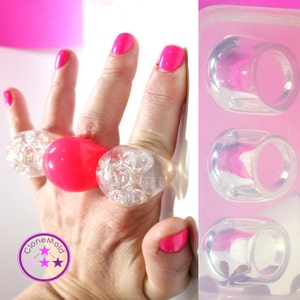 Puede incluir: Un molde de silicona para hacer anillos de burbujas. El molde tiene tres cavidades, cada una con forma de anillo con un diseño de burbujas. El molde se muestra junto a una mano que lleva tres anillos hechos con el molde.