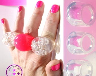 May include: A silicone mold for making bubble rings. The mold has three cavities, each shaped like a ring with a bubble design. The mold is shown next to a hand wearing three rings made with the mold.