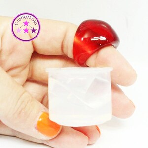May include: A large, red, domed ring is worn on a finger. A small, translucent, white container is held in the same hand. The ring has a smooth, glossy finish. The container is cylindrical with a slightly flared top. The image includes the text "cloneMold".