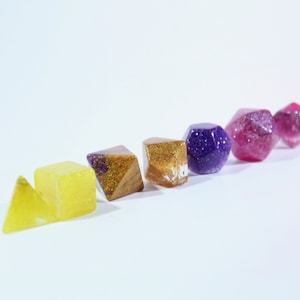 May include: A set of six resin dice in various shapes and colors, including yellow, gold, purple, and pink. The dice have a glittery finish.