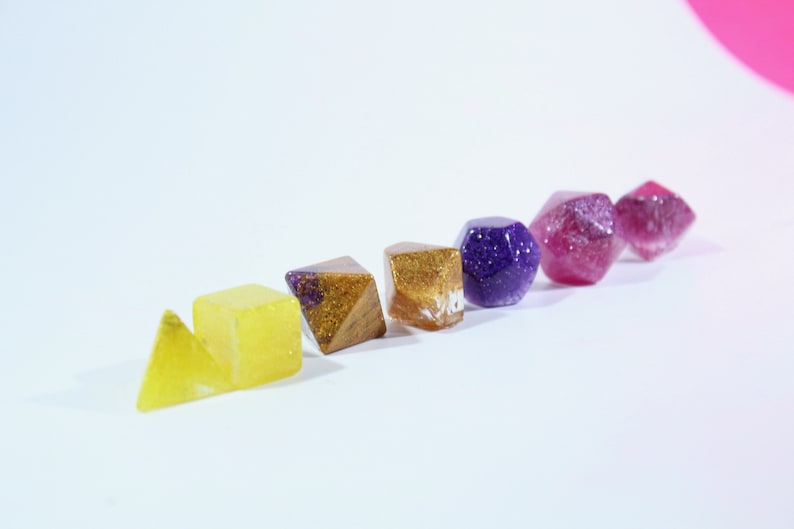 May include: A set of six resin dice in various shapes and colors, including yellow, gold, purple, and pink. The dice have a glittery finish.
