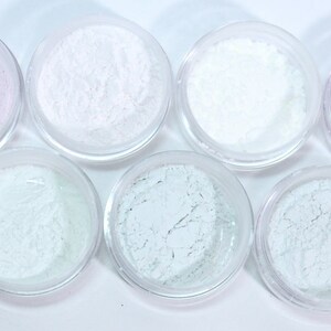 May include: Seven small, clear jars filled with various shades of powder. The colors include white, pale lavender, and several shades of pale green. The powders have a fine, textured appearance, and the jars are arranged in a circular pattern.